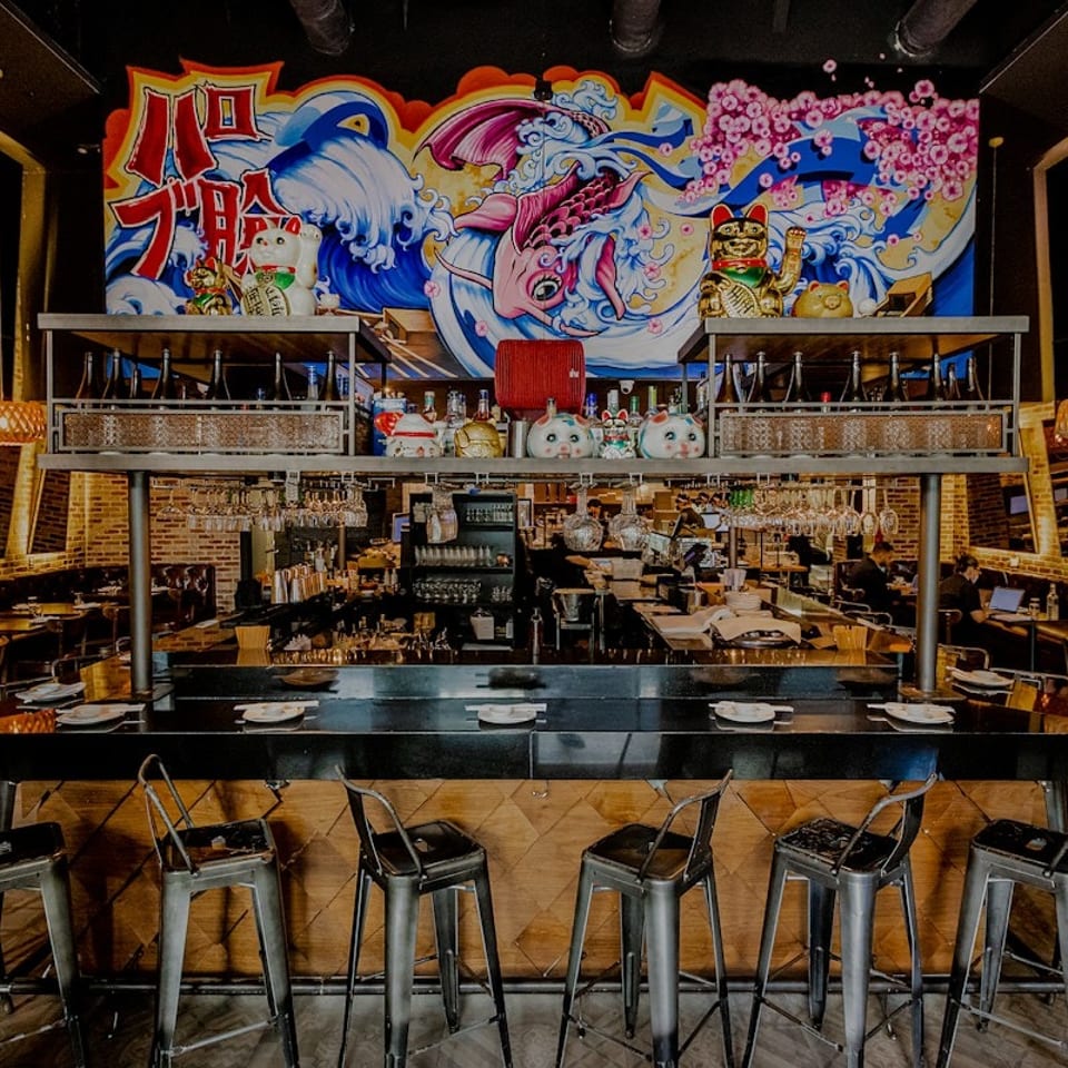 Modern restaurant interior with a long bar, high stools, and a vibrant Japanese-style mural.