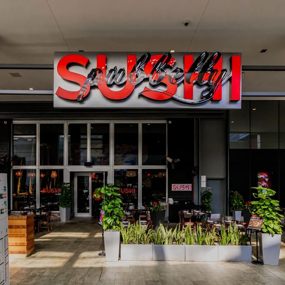 The exterior of a modern sushi restaurant with a large red "Sushi" sign and outdoor seating.