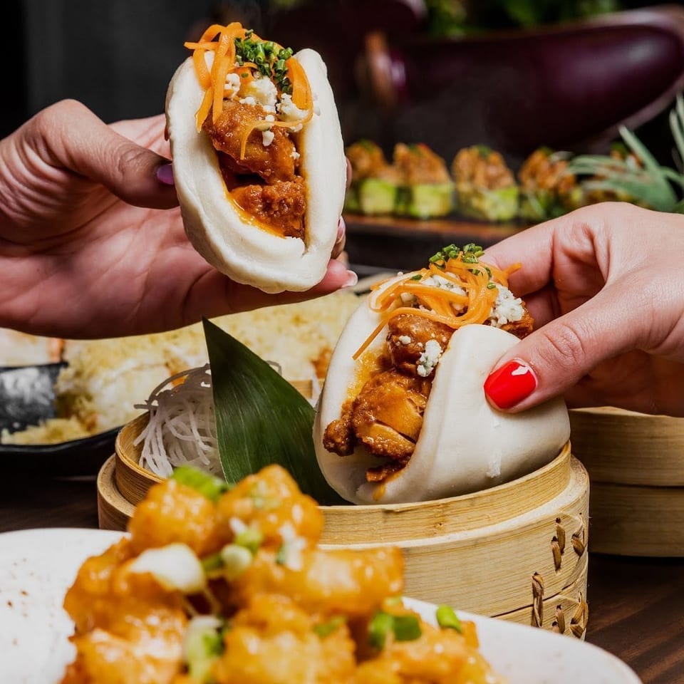 Two hands holding fluffy bao buns filled with chicken, carrots, and cheese, with other dishes blurred.