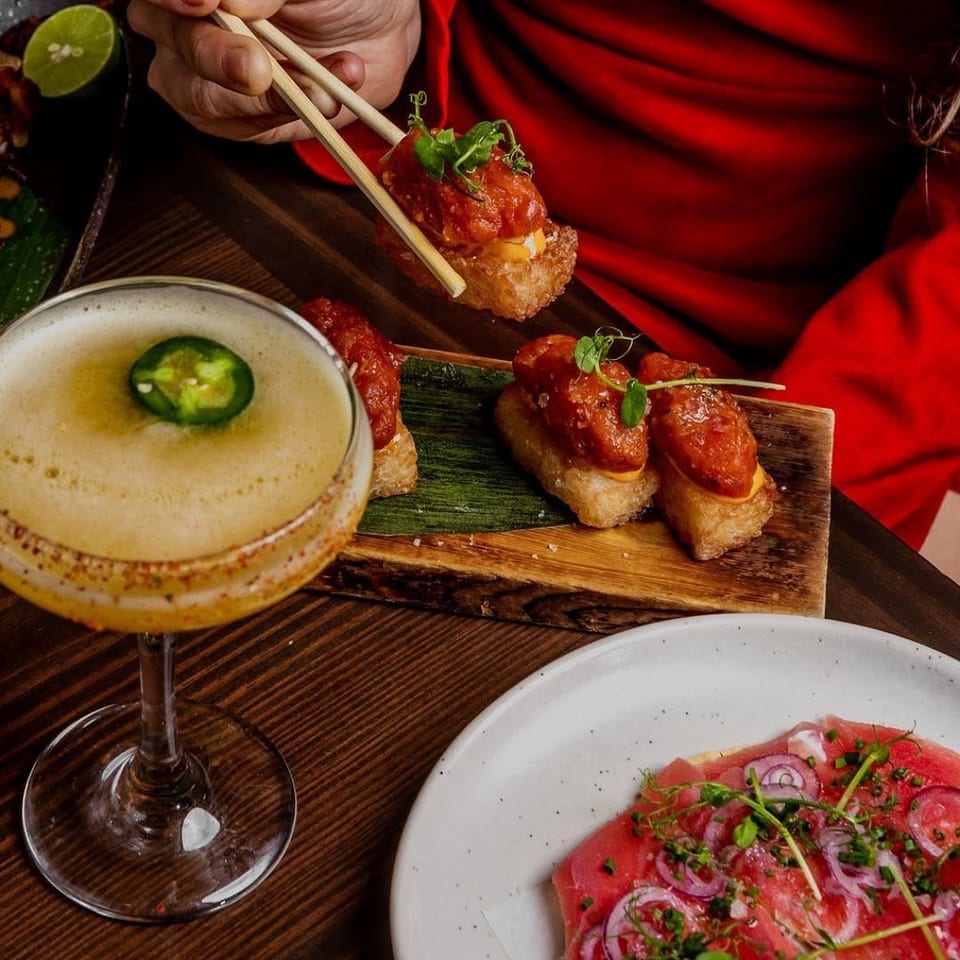 Chopsticks holding a piece of spicy tuna crispy rice, with a cocktail and tuna carpaccio.